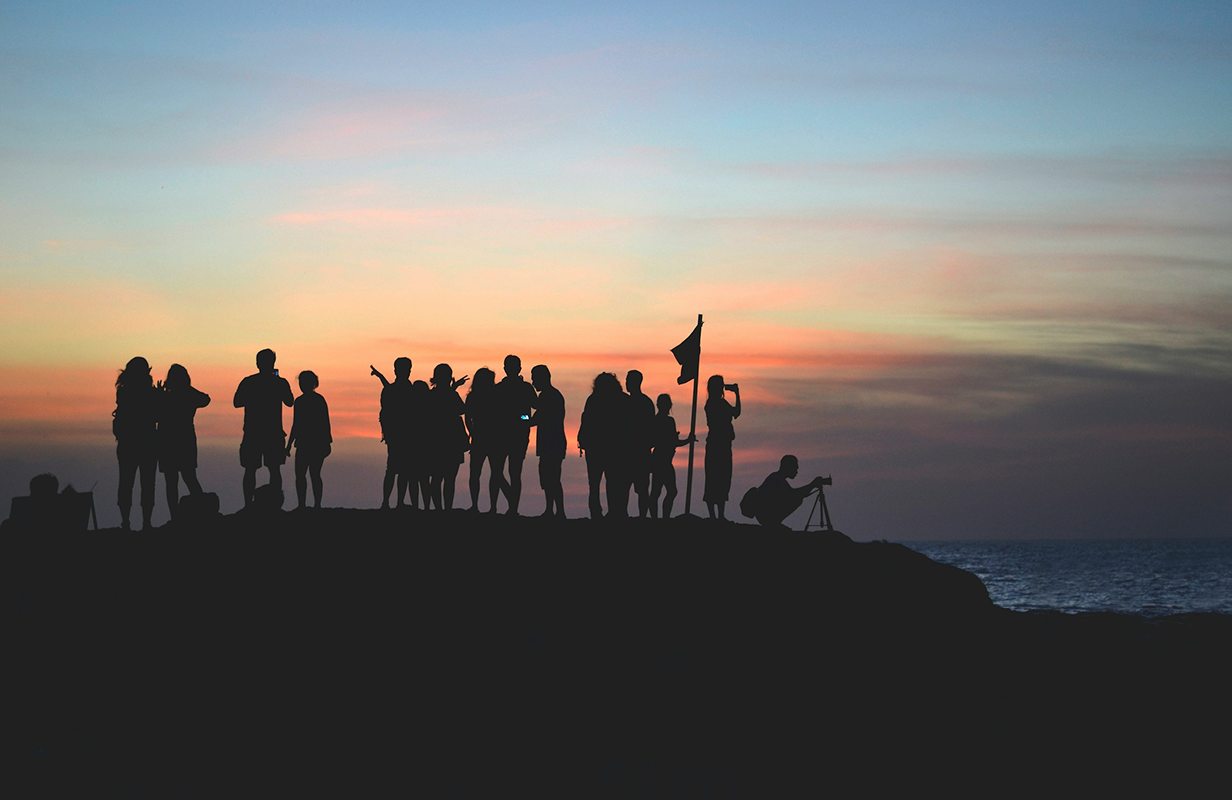 Symbolbild: Eine Gruppe von Menschen ist in Silhouette auf einem Hügel vor einem Sonnenuntergang zu sehen. Sie stehen vor einem ruhigen Meer. Am rechten Rand der Gruppe kniet eine Person mit einer Kamera auf einem Stativ.