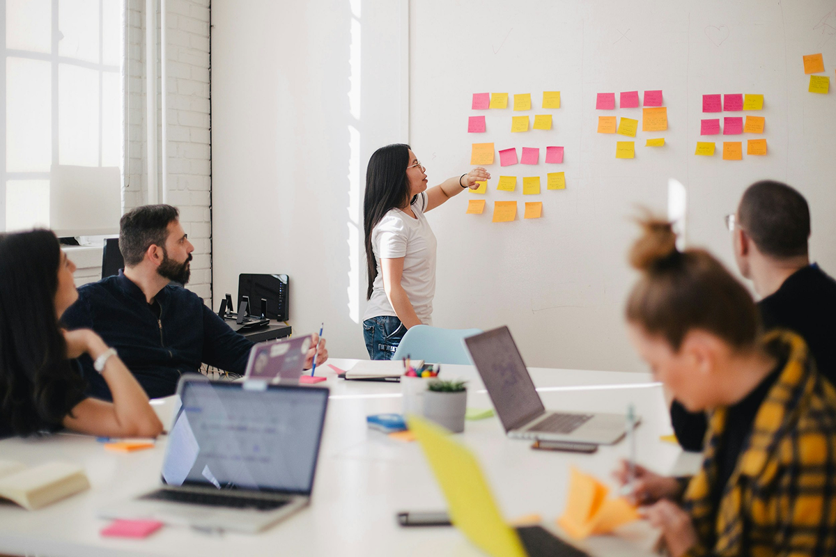 Stock photo: A woman in an office stands in front of a white wall, pointing at yellow and pink sticky notes. Her team sits around a conference table with laptops, watching her.